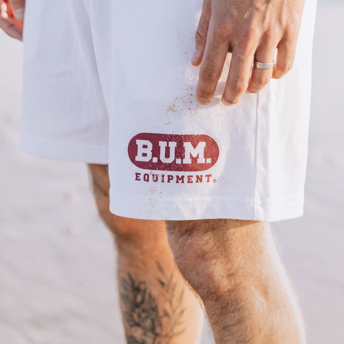 Close crop of the B.U.M. Mens Shorts in white on the beach, a hand brushing sand off the leg beside the red B.U.M. Equipment badge