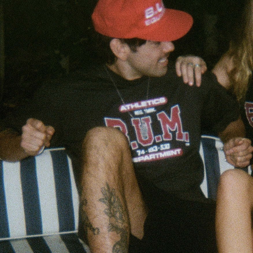 Man on a striped bench at night wearing the B.U.M. Mens T-Shirt in black with the ATHLETICS DEPARTMENT print and a red B.U.M. Equipment cap