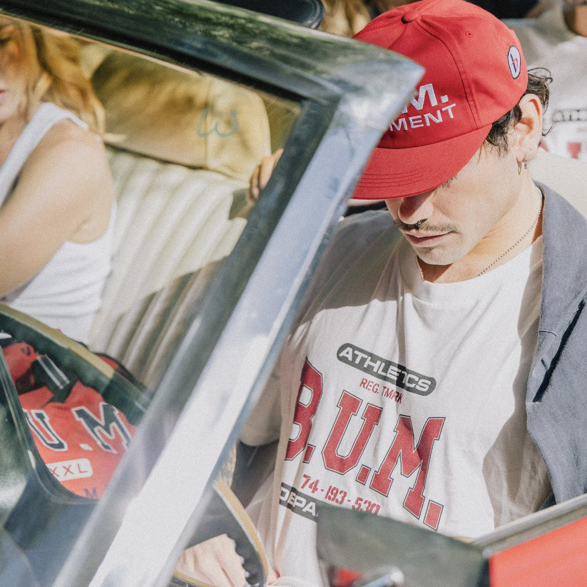 Man in a red B.U.M. Equipment cap wearing the B.U.M. T-Shirt in white, leaning on a car