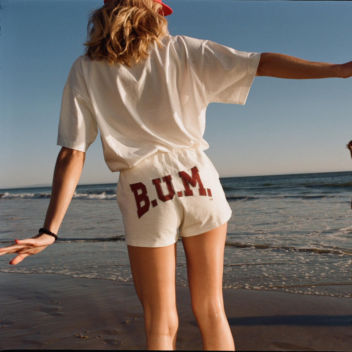 Woman on the beach in the B.U.M. Womens Shorts Off-White with a white B.U.M. tee, seen from behind with the arched B.U.M. logo across the back of the shorts